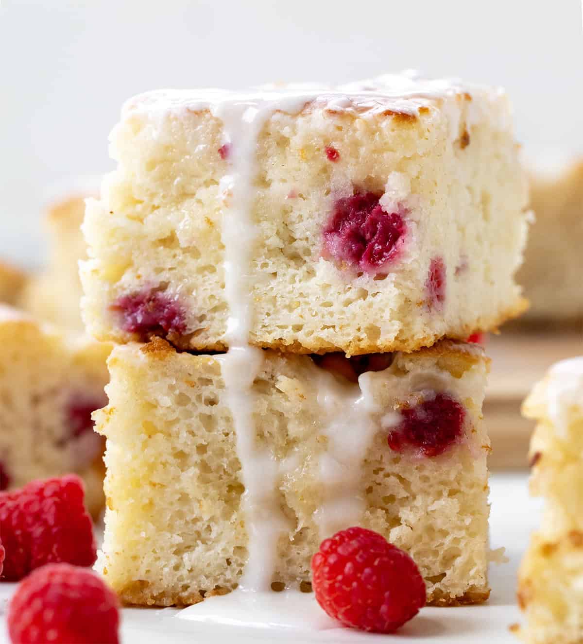 Soft, buttery Glazed Raspberry Butter Swim Biscuits are filled with juicy raspberries and baked right in melted butter for golden, crispy edges and a tender center. Topped with a sweet glaze, they are an easy, bakery-style treat made with simple ingredients with no rolling or cutting needed! Stack of Raspberry Butter Swim Biscuits on a white table surrounded by other biscuits and fresh raspberries.