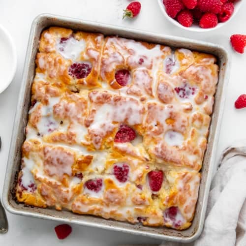 Pan of glazed Raspberry Butter Swim Biscuits on a white table with fresh raspberries from overhead.