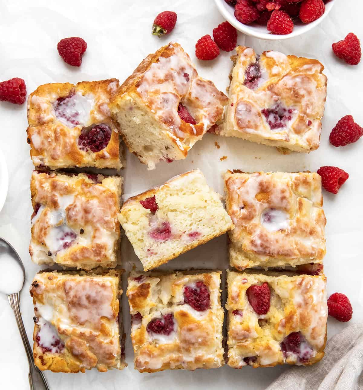 Soft, buttery Glazed Raspberry Butter Swim Biscuits are filled with juicy raspberries and baked right in melted butter for golden, crispy edges and a tender center. Topped with a sweet glaze, they are an easy, bakery-style treat made with simple ingredients with no rolling or cutting needed! Glazed Raspberry Butter Swim Biscuits cut into squares and surrounded by raspberries.