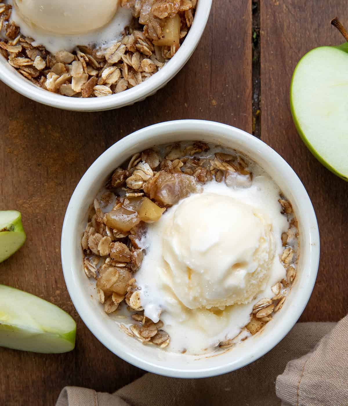 Craving a quick and satisfying dessert? This apple crisp is the perfect single-serve treat. Made with fresh apples and a simple brown sugar oat topping, it comes together in minutes, no oven needed! Single serve bowls of Apple Crisp on a wooden table with forks and fresh green apples.