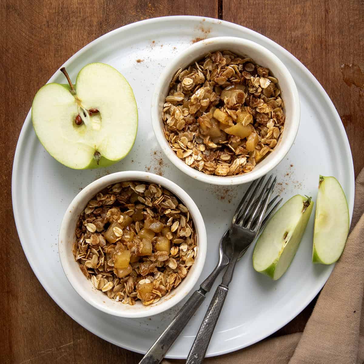 Craving a quick and satisfying dessert? This apple crisp is the perfect single-serve treat. Made with fresh apples and a simple brown sugar oat topping, it comes together in minutes, no oven needed! Two Single Serve Apple Crisp's on a white round platter on a wooden table from overhead.