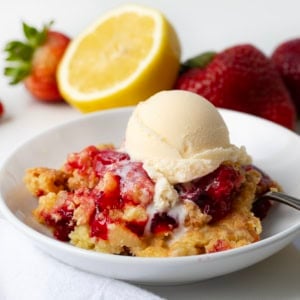 White plate of Strawberry Lemon Dump Cake on a white table with fresh lemon and strawberries in the background.