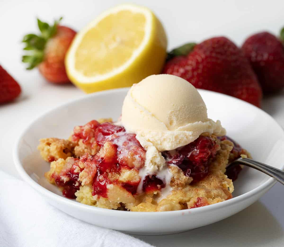 If you are looking for an easy, like really easy dessert, this Strawberry Lemon Dump Cake is it! With just four ingredients, you get a bright strawberry lemon flavor in every bite. Serve it warm with a scoop of vanilla ice cream or a dollop of whipped cream. Or, just grab a spoon and dig in as-is! White plate of Strawberry Lemon Dump Cake on a white table with fresh lemon and strawberries in the background.