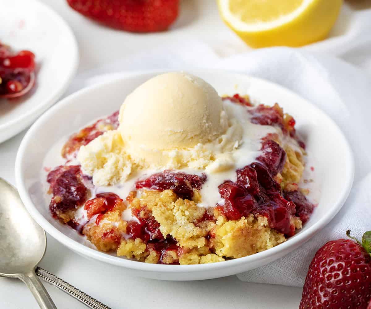 If you are looking for an easy, like really easy dessert, this Strawberry Lemon Dump Cake is it! With just four ingredients, you get a bright strawberry lemon flavor in every bite. Serve it warm with a scoop of vanilla ice cream or a dollop of whipped cream. Or, just grab a spoon and dig in as-is! White plate of Strawberry Lemon Dump Cake on a white table with fresh lemon and strawberries in the background.
