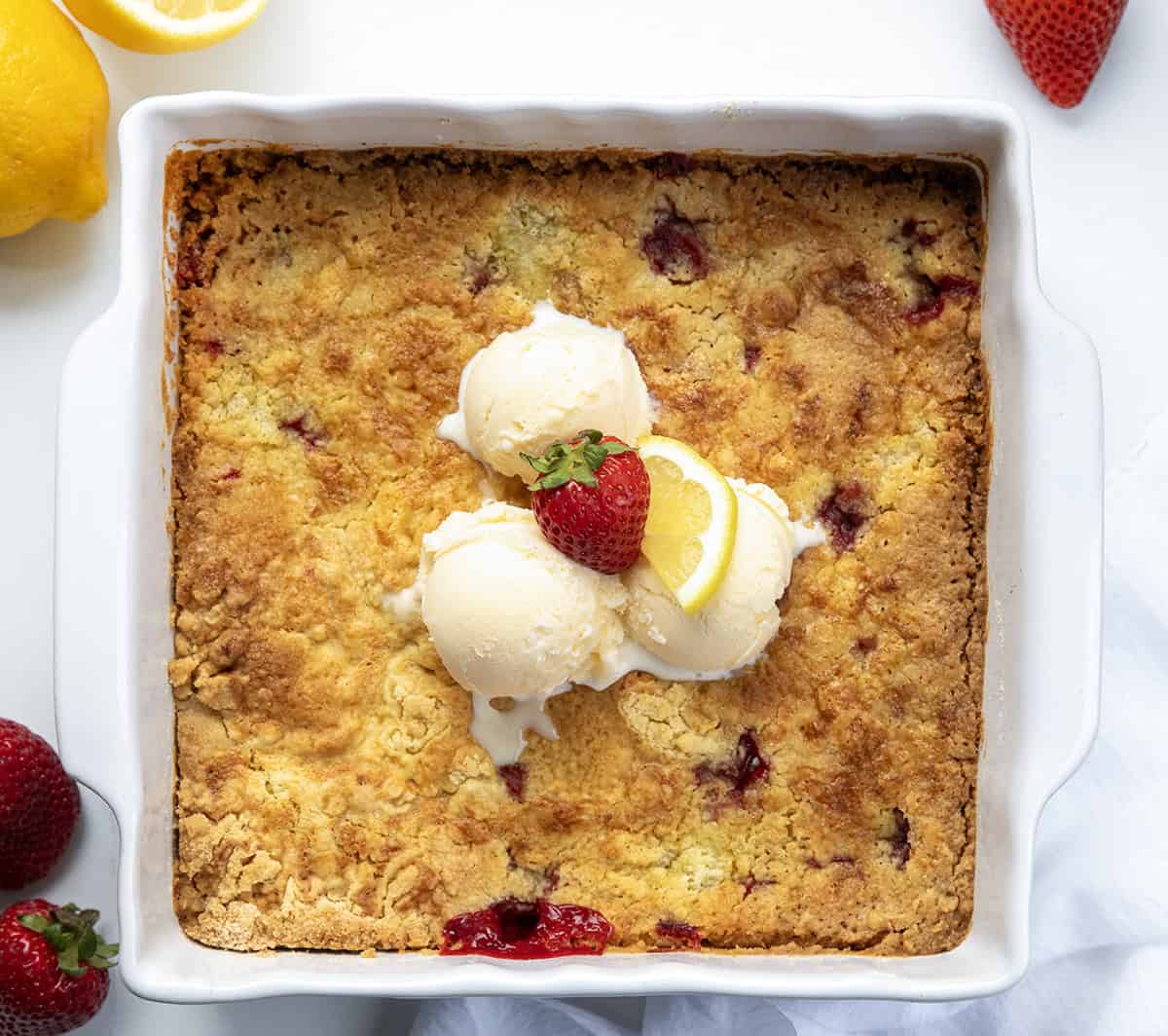 If you are looking for an easy, like really easy dessert, this Strawberry Lemon Dump Cake is it! With just four ingredients, you get a bright strawberry lemon flavor in every bite. Serve it warm with a scoop of vanilla ice cream or a dollop of whipped cream. Or, just grab a spoon and dig in as-is! Pan of Strawberry Lemon Dump Cake on a white table with ice cream and strawberries from overhead.