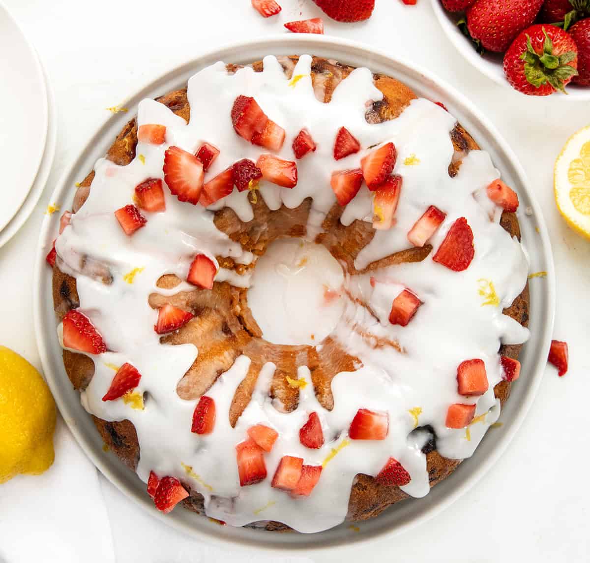 Strawberry Lemon Pound Cake is a moist and buttery homemade pound cake filled with fresh strawberries, bright lemon flavor, and topped with a tangy lemon glaze. Itโs the perfect spring or summer dessert! Whole Strawberry Lemon Pound Cake on a white cake plate on a white table from overhead.