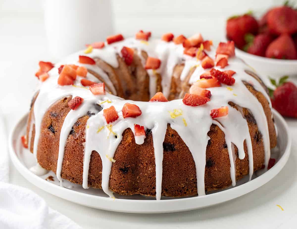 Strawberry Lemon Pound Cake is a moist and buttery homemade pound cake filled with fresh strawberries, bright lemon flavor, and topped with a tangy lemon glaze. Itโs the perfect spring or summer dessert! Whole Strawberry Lemon Pound Cake on a white cake plate on a white table close up.