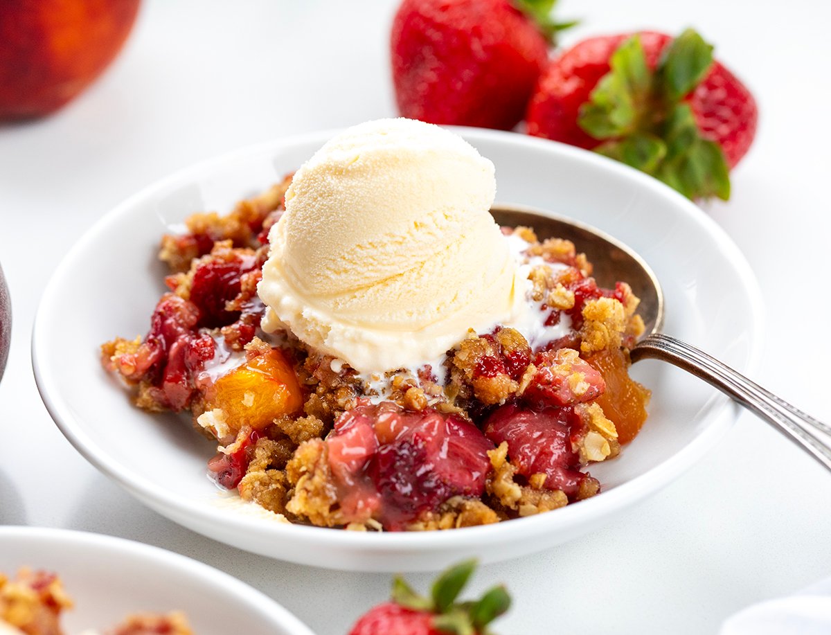 This Strawberry Peach Crisp is the perfect summer dessert, made with juicy, sweet strawberries and ripe peaches baked under a buttery oat topping with warm cinnamon and brown sugar. Every bite is bursting with fruit flavor and a crumbly texture. I love how easy this recipe is to throw together! Bowl of Strawberry Peach Crisp on a white table with a spoon in the bowl.