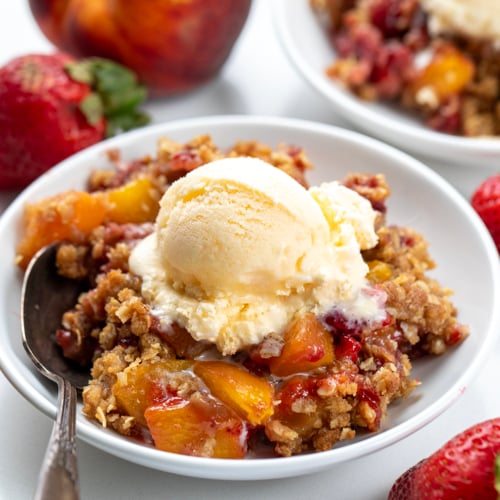 White bowls filled with Strawberry Peach Crisp on a white table with fresh peaches and strawberries.