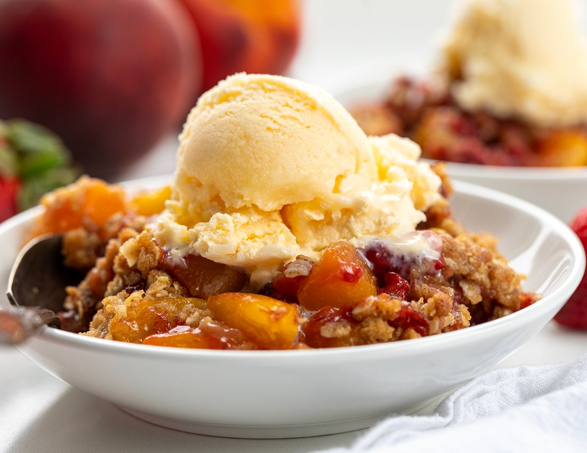 This Strawberry Peach Crisp is the perfect summer dessert, made with juicy, sweet strawberries and ripe peaches baked under a buttery oat topping with warm cinnamon and brown sugar. Every bite is bursting with fruit flavor and a crumbly texture. I love how easy this recipe is to throw together! Close up of a bowl of Strawberry Peach Crisp with a scoop of vanilla ice cream on it.