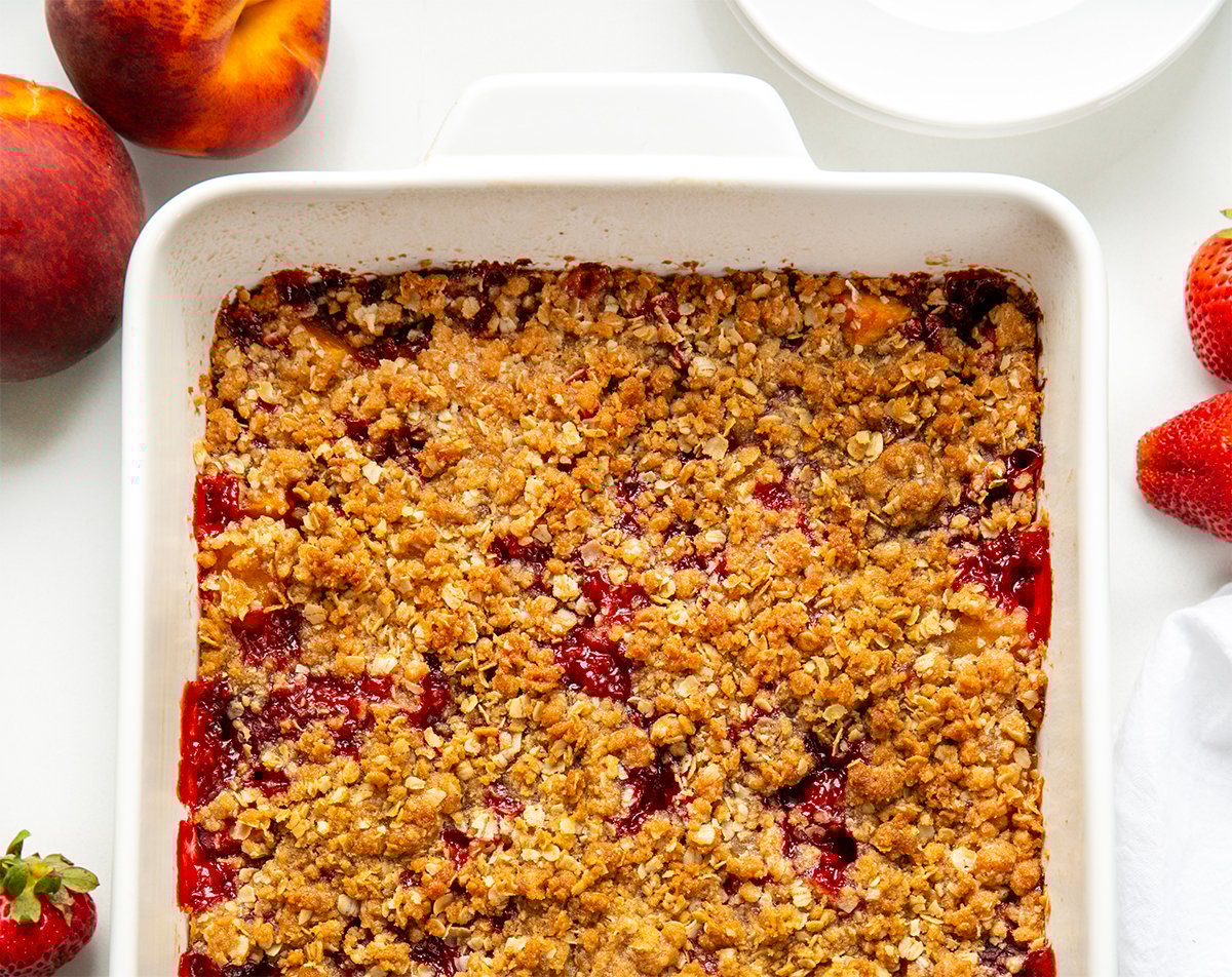 This Strawberry Peach Crisp is the perfect summer dessert, made with juicy, sweet strawberries and ripe peaches baked under a buttery oat topping with warm cinnamon and brown sugar. Every bite is bursting with fruit flavor and a crumbly texture. I love how easy this recipe is to throw together! Pan of Strawberry Peach Crisp on a white table from overhead.