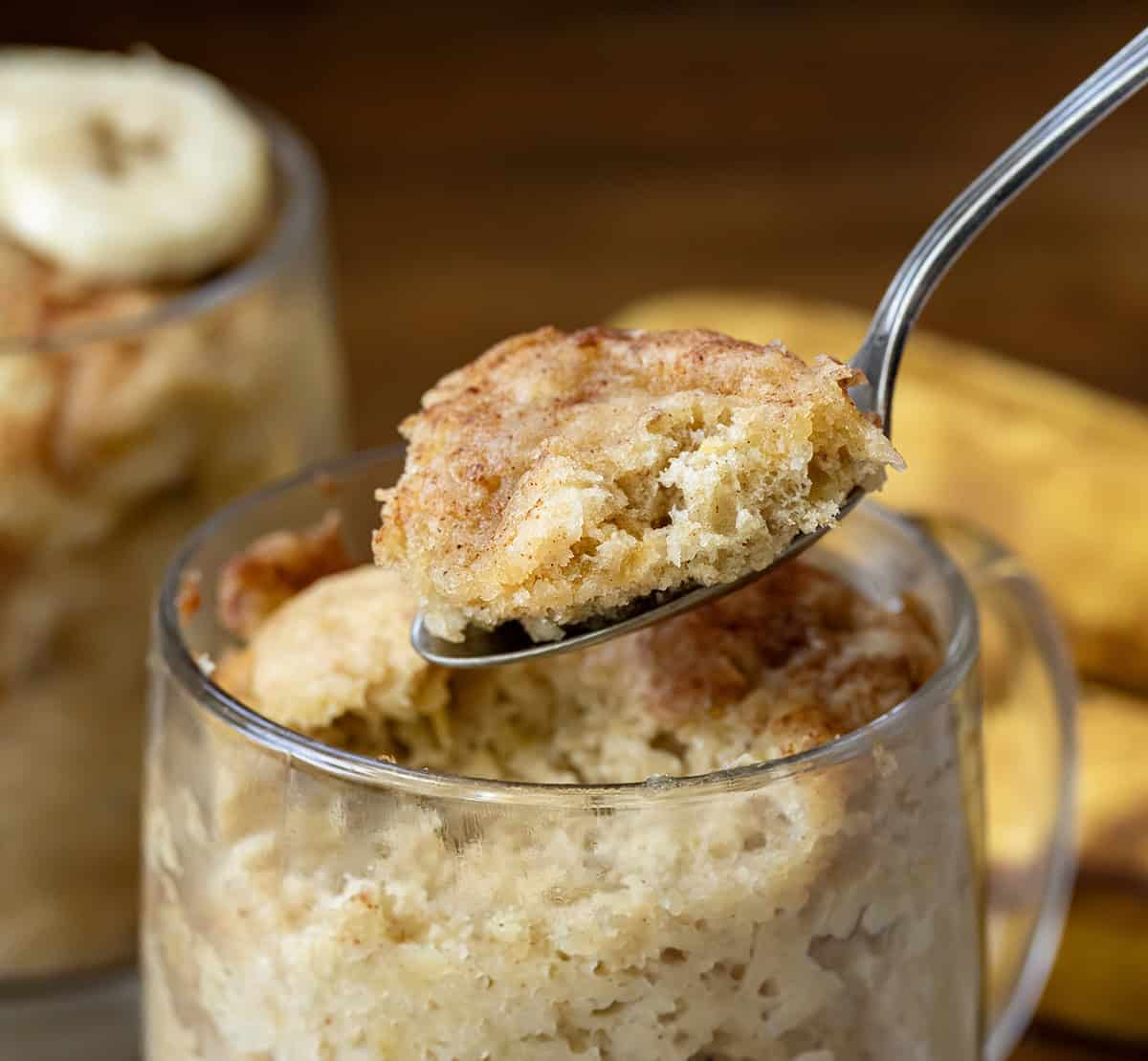 This microwave Banana Mug Cake is a quick and easy way to enjoy all the classic flavors of banana bread, packed with plenty of banana goodness and a warm touch of cinnamon. Ready in just minutes, it’s the perfect cozy dessert without any fuss! Spoonful of Banana Cake out of a mug on a wooden table.