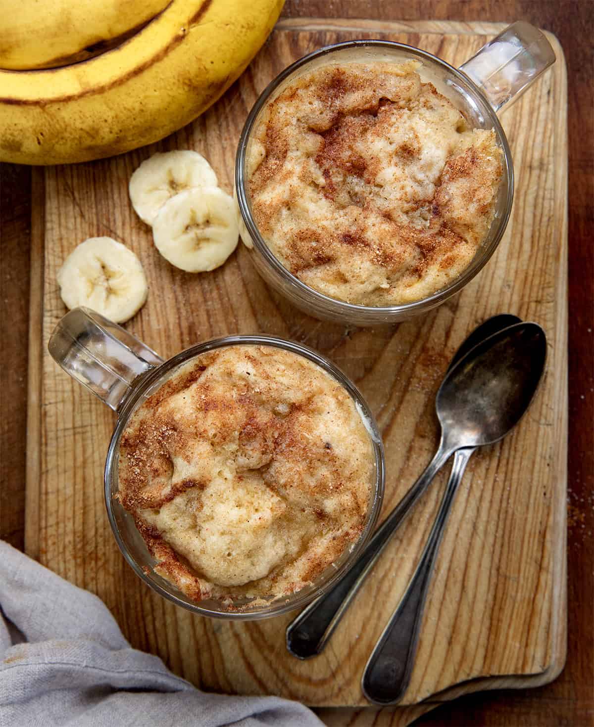 This microwave Banana Mug Cake is a quick and easy way to enjoy all the classic flavors of banana bread, packed with plenty of banana goodness and a warm touch of cinnamon. Ready in just minutes, it’s the perfect cozy dessert without any fuss! Banana Mug Cakes on a wooden table from overhead.