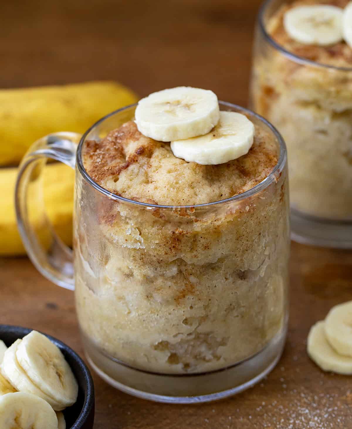 This microwave Banana Mug Cake is a quick and easy way to enjoy all the classic flavors of banana bread, packed with plenty of banana goodness and a warm touch of cinnamon. Ready in just minutes, it’s the perfect cozy dessert without any fuss! Banana Mug Cakes on a wooden table with bananas.