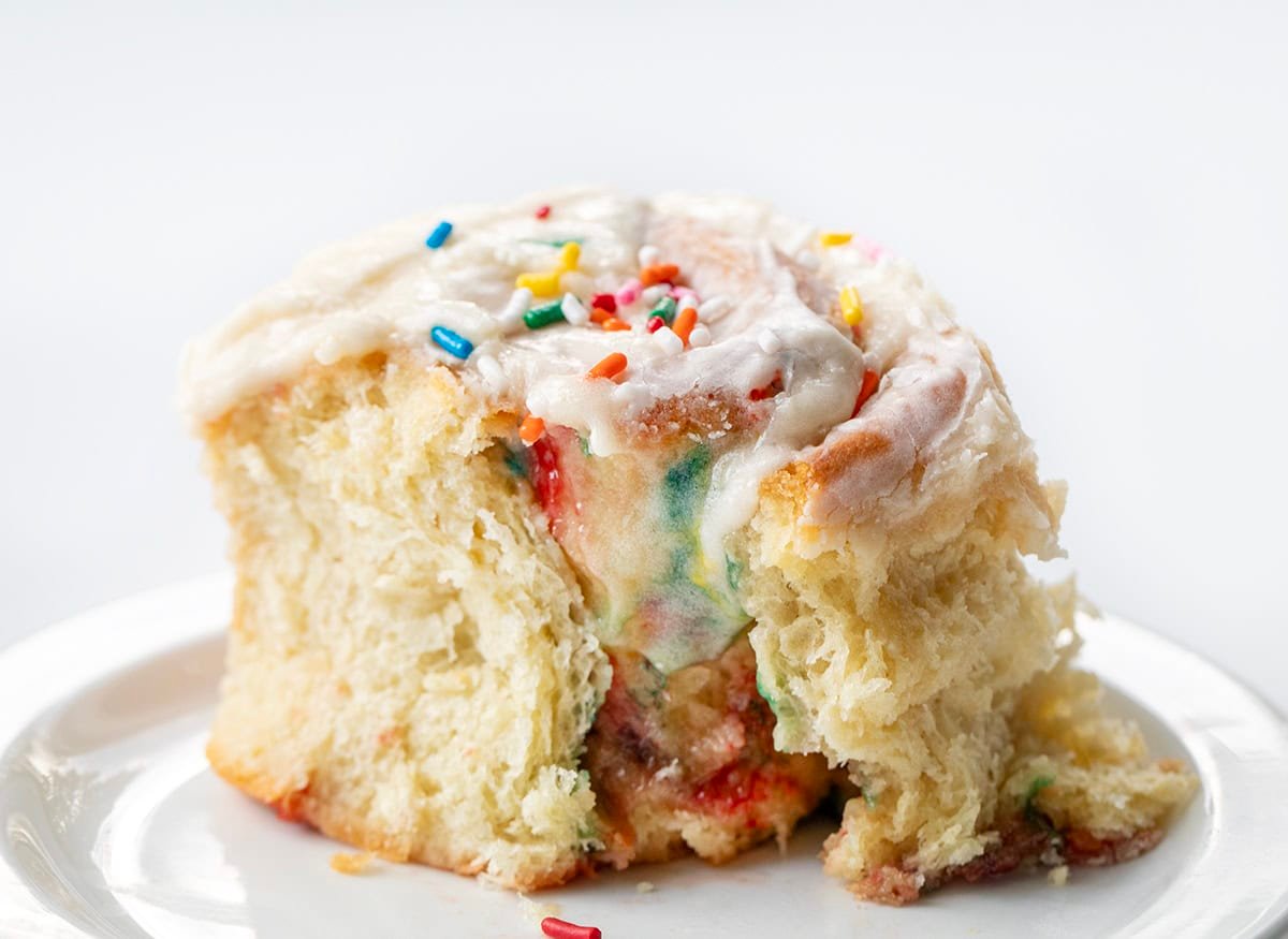 Celebrate every day like it's your birthday with these Birthday Cake Cinnamon Rolls! Soft, fluffy, mile-high rolls, like my classic cinnamon rolls, are filled with a sweet, buttery brown sugar mixture and loaded with colorful rainbow sprinkles that bring fun and festive flavor to every bite! Birthday Cake Cinnamon Roll on a plate with some of the roll removed showing the melting frosting inside.