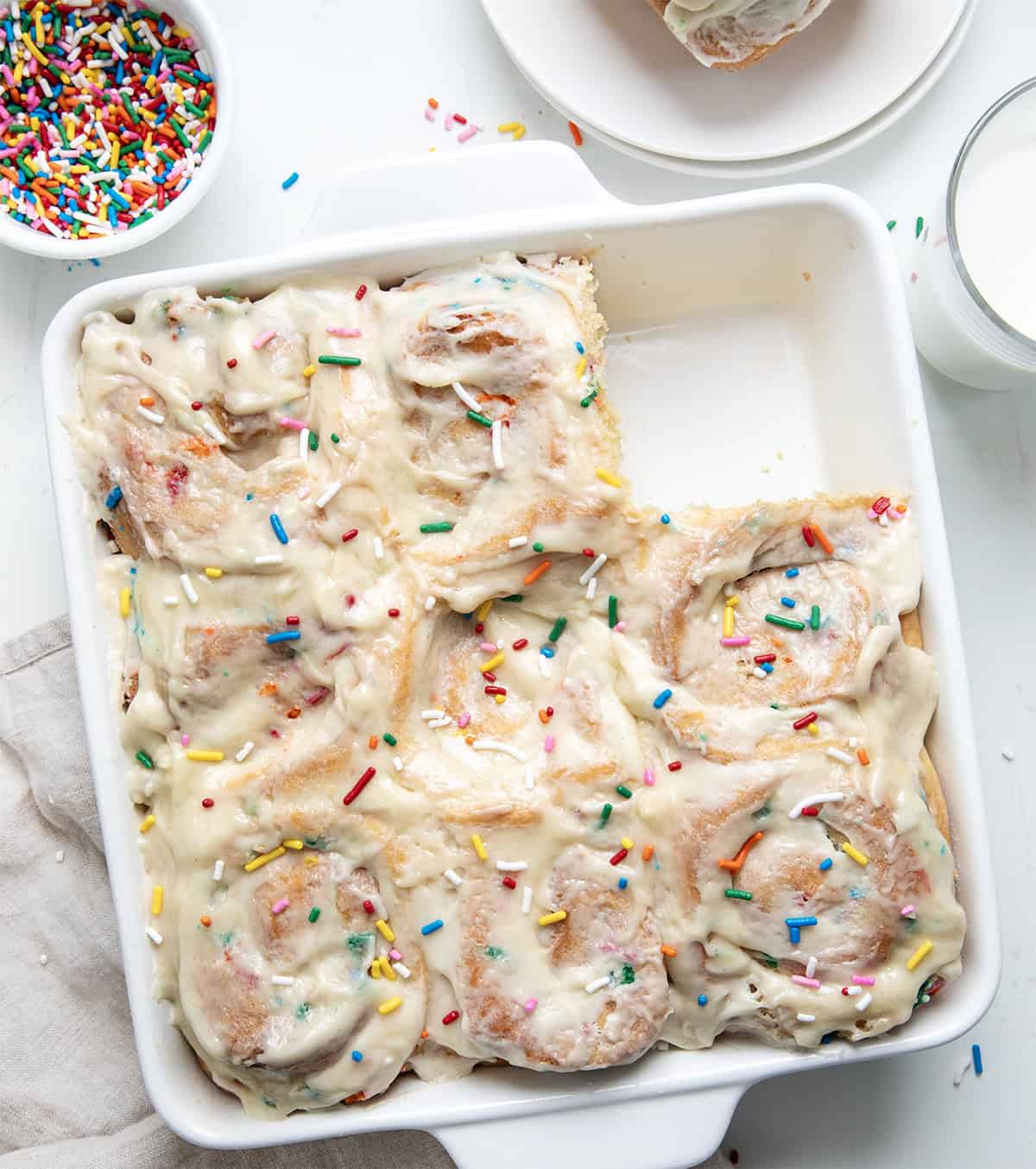 Celebrate every day like it's your birthday with these Birthday Cake Cinnamon Rolls! Soft, fluffy, mile-high rolls, like my classic cinnamon rolls, are filled with a sweet, buttery brown sugar mixture and loaded with colorful rainbow sprinkles that bring fun and festive flavor to every bite! Pan of Birthday Cake Cinnamon Rolls with one missing on a white table from overhead.