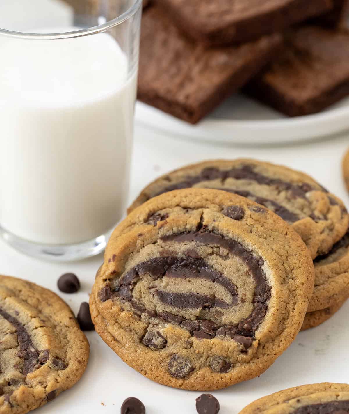 These semi-homemade Brownie Cookie Swirls combine classic chocolate chip dough with rich, fudgy brownie batter in one delicious treat! They are soft, chewy, full of chocolatey goodness, and surprisingly easy to make with just a handful of ingredients! Brownie Cookie Swirls on a white table with milk and brownies in the background.