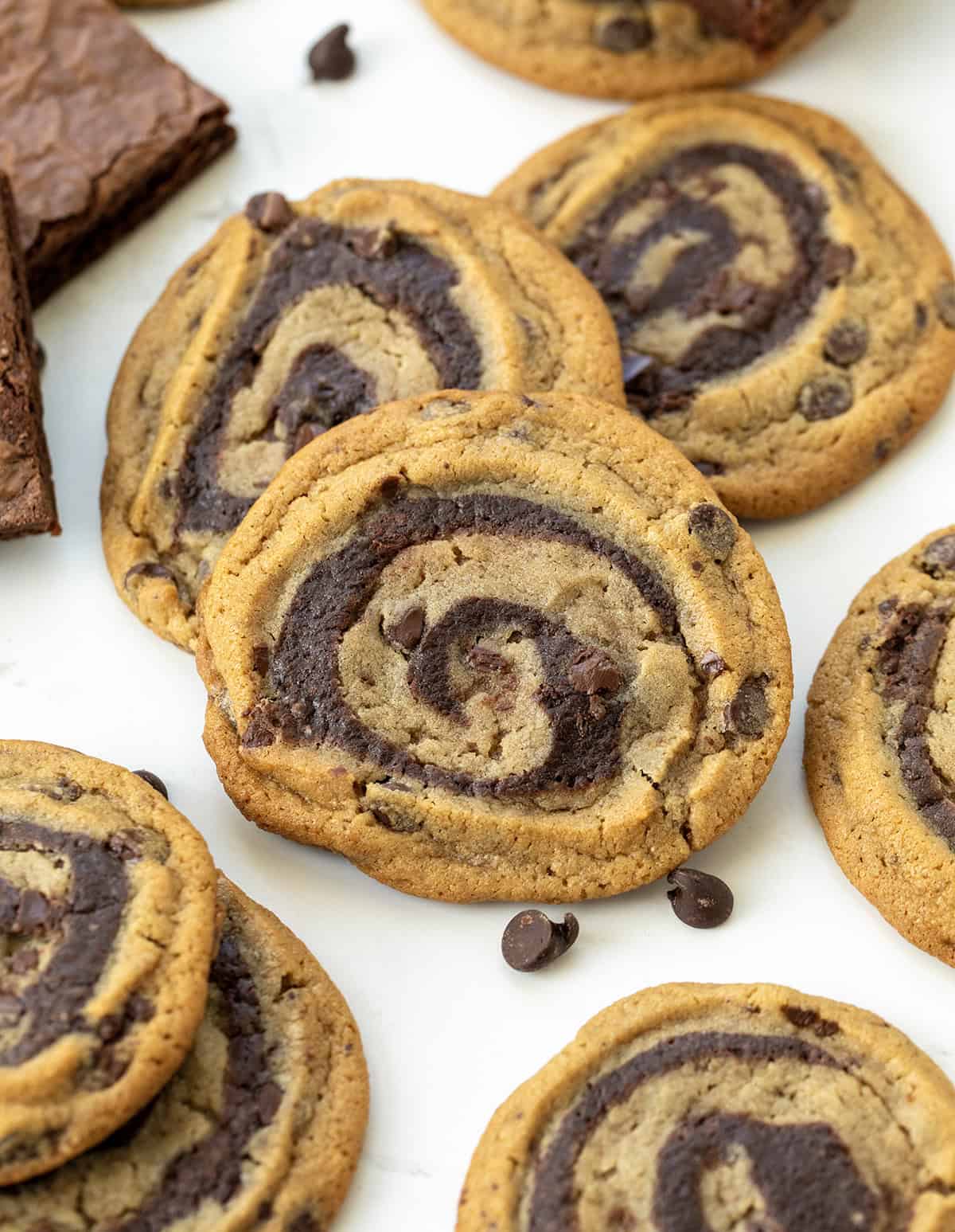 These semi-homemade Brownie Cookie Swirls combine classic chocolate chip dough with rich, fudgy brownie batter in one delicious treat! They are soft, chewy, full of chocolatey goodness, and surprisingly easy to make with just a handful of ingredients! Close up of lots of Brownie Cookie Swirls on a white table with chocolate morsels and brownies off to the side.