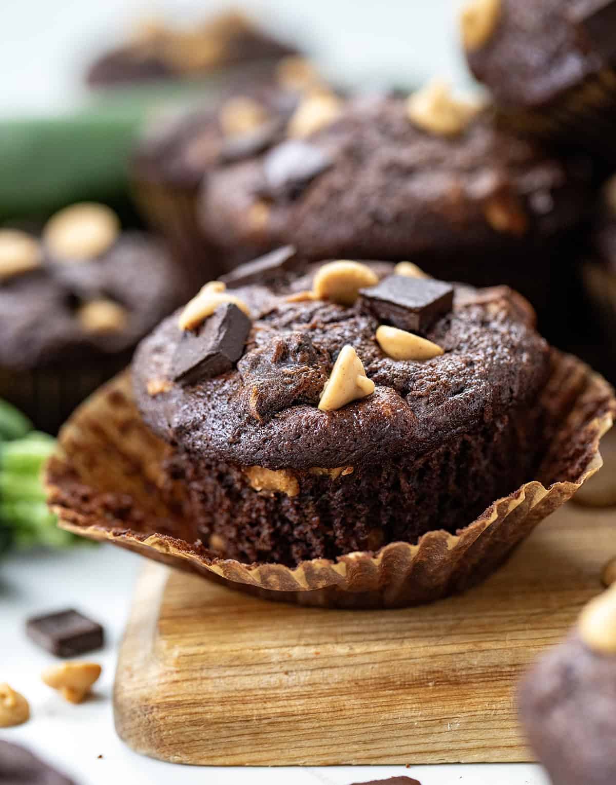 Chocolate Peanut Butter Zucchini Muffin with its wrapper pulled down on a cutting board surrounded by more muffins and fresh zucchini.