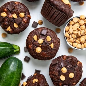 Chocolate Peanut Butter Zucchini Muffins on a white table with fresh zucchini and peanut butter chips nearby.
