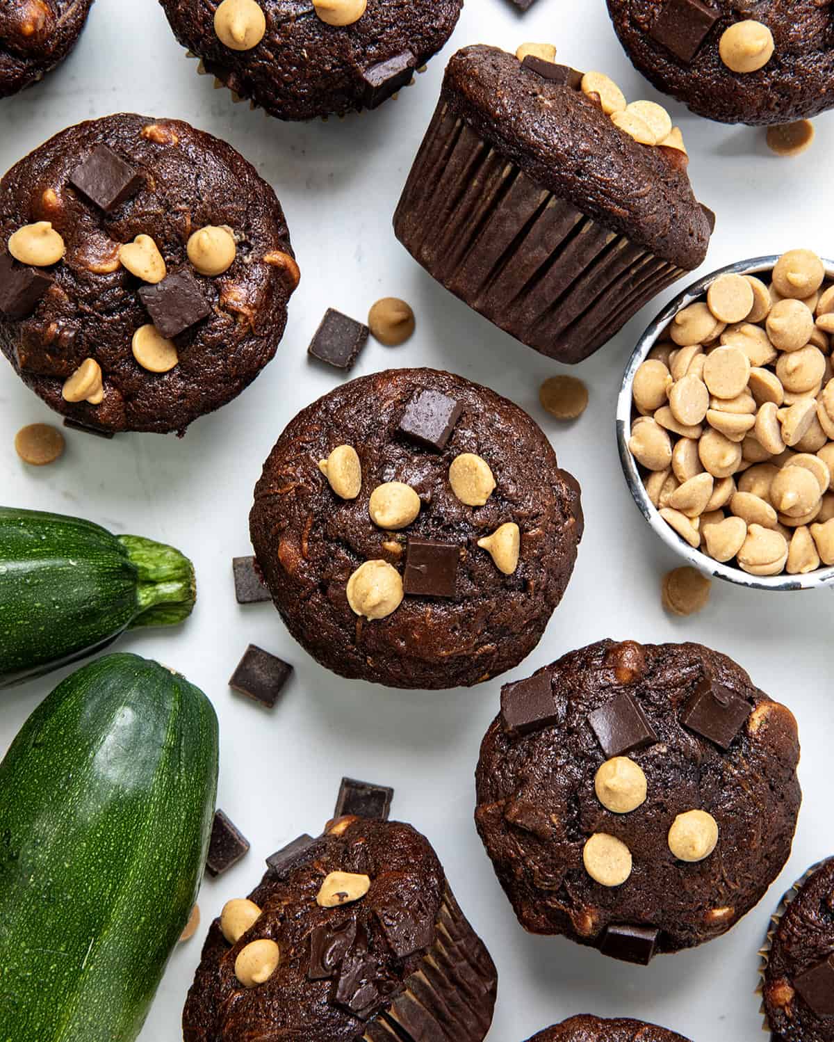 Chocolate Peanut Butter Zucchini Muffins on a white table with fresh zucchini and peanut butter chips nearby.