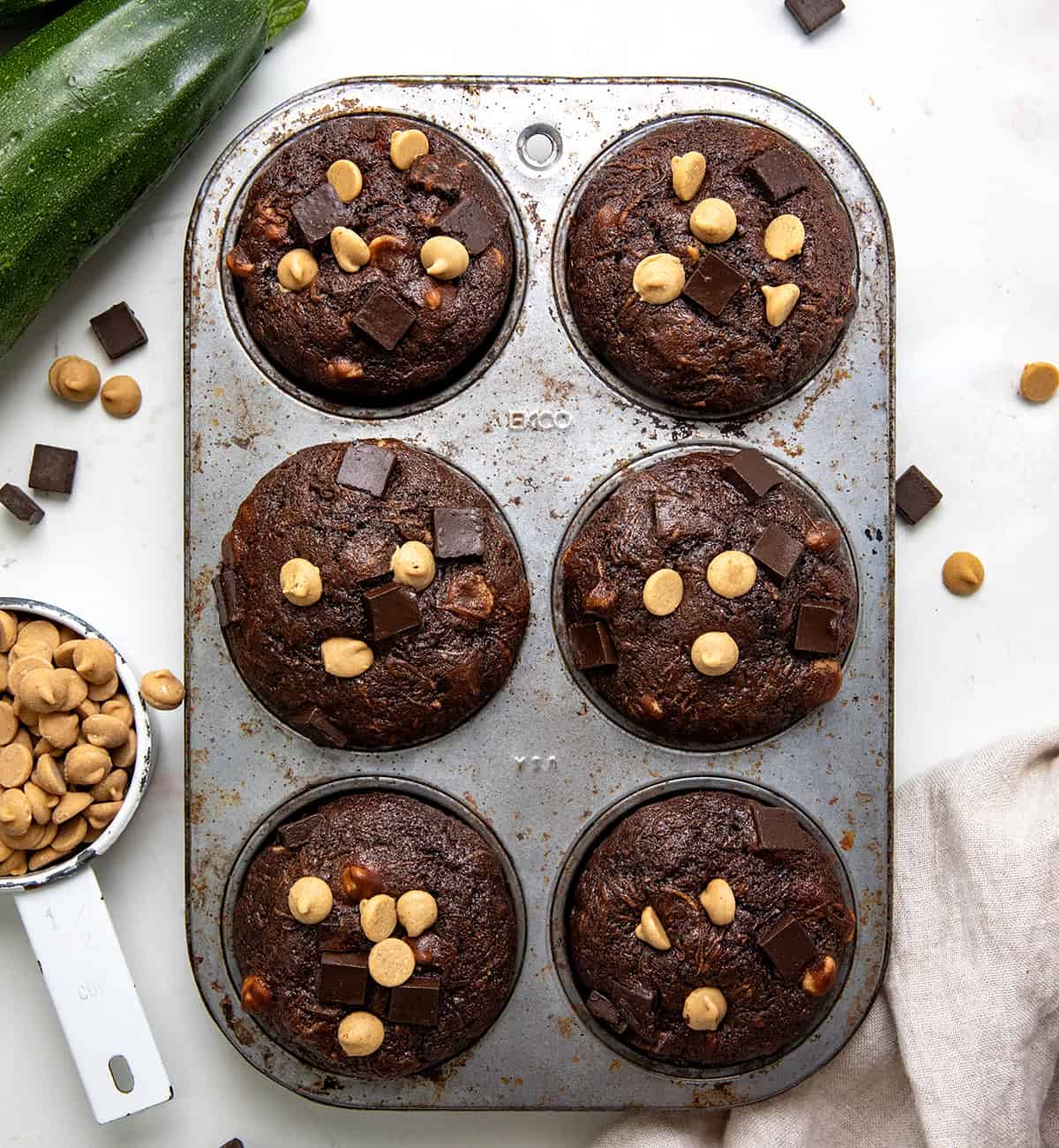 Pan of Chocolate Peanut Butter Zucchini Muffins on a white table with chocolate, peanut butter chips, and zucchini nearby.