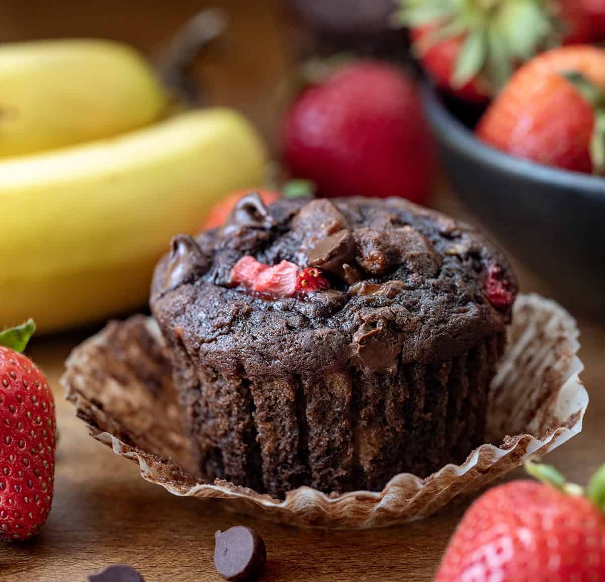 If you love the combination of fruit and chocolate, these Chocolate Strawberry Banana Muffins are about to become your new favorite banana muffin recipe! Rich, moist, and packed with flavor, this easy muffin recipe combines ripe bananas, melty chocolate chips, and bursts of strawberry in every bite! Chocolate Strawberry Banana Muffins with its wrapped pulled down on a wooden table with fresh strawberries and banana.