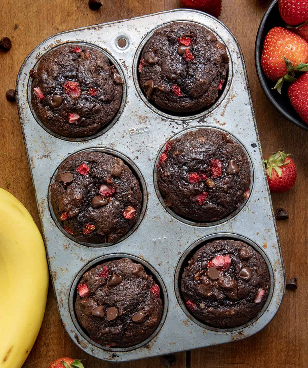 If you love the combination of fruit and chocolate, these Chocolate Strawberry Banana Muffins are about to become your new favorite banana muffin recipe! Rich, moist, and packed with flavor, this easy muffin recipe combines ripe bananas, melty chocolate chips, and bursts of strawberry in every bite! Chocolate Strawberry Banana Muffins in a muffin pan on a wooden tale from overhead.