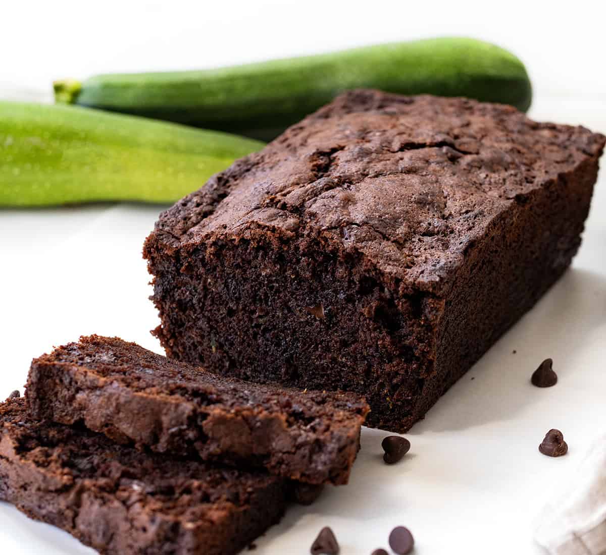 This Chocolate Zucchini Bread is rich, moist, and loaded with melty chocolate chips! Proof that veggies and dessert really can be best friends. With deep cocoa flavor, a perfectly tender crumb, and just the right amount of sweetness, you’d never guess there’s zucchini hiding inside! Loaf of Chocolate Zucchini Bread with a couple of pieces sliced off on a white table.