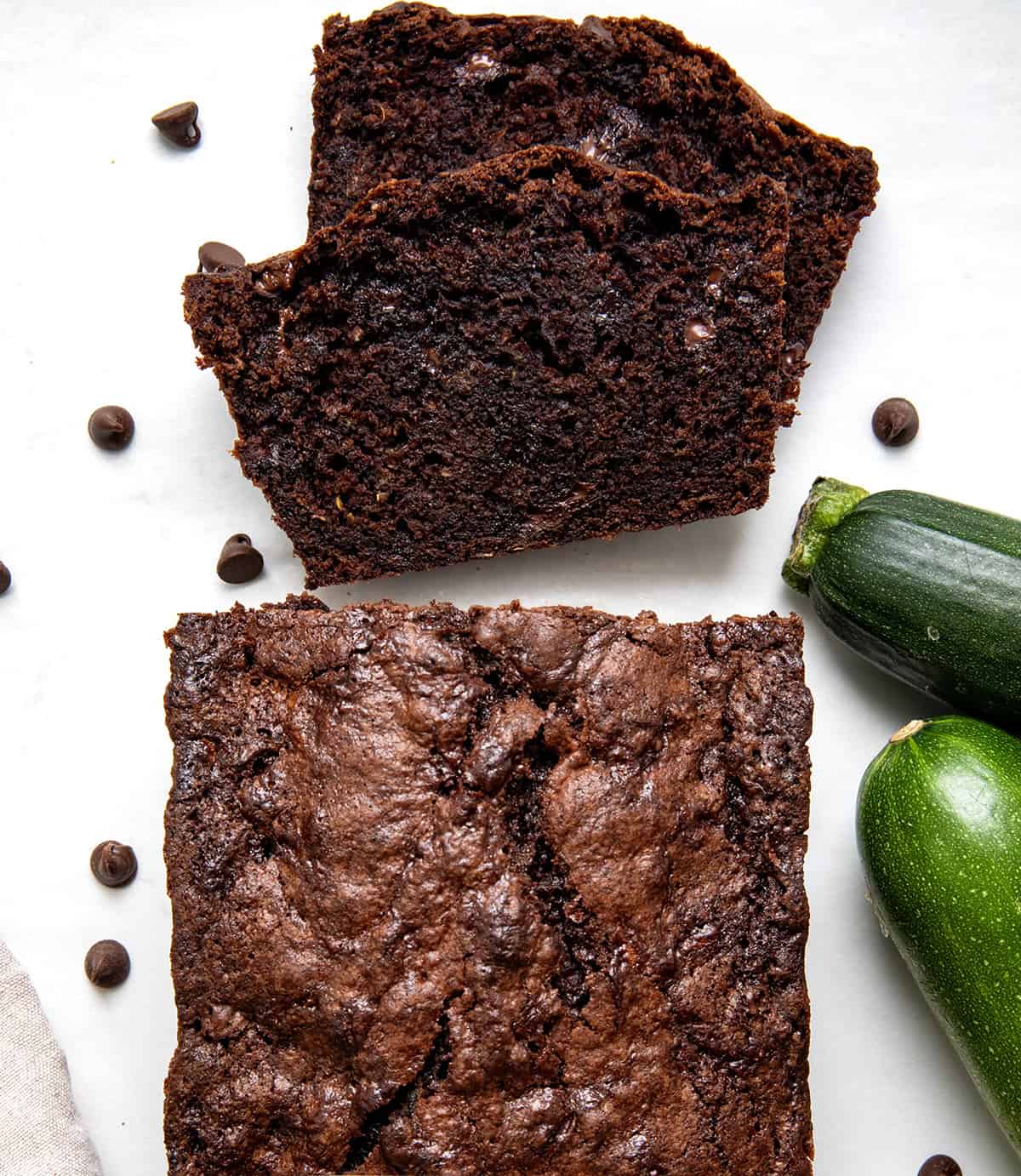 This Chocolate Zucchini Bread is rich, moist, and loaded with melty chocolate chips! Proof that veggies and dessert really can be best friends. With deep cocoa flavor, a perfectly tender crumb, and just the right amount of sweetness, you’d never guess there’s zucchini hiding inside! Loaf of Chocolate Zucchini Bread on a white table with a couple of pieces cut and laying flat.