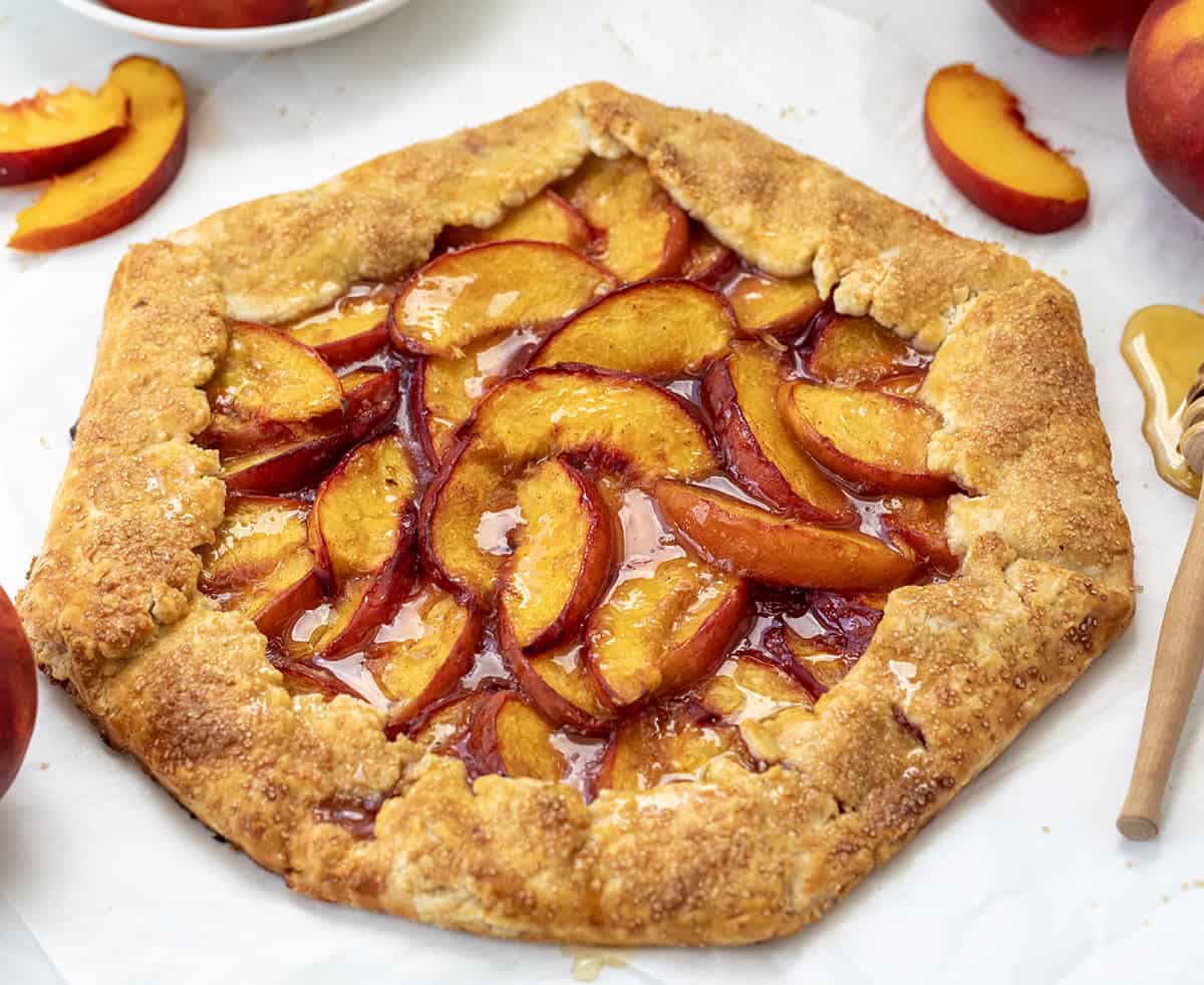 This easy Peach Galette is the perfect summer dessert, made with juicy, fresh peaches wrapped in a buttery, flaky pie crust and baked until golden and bubbling. It is simpler than pie, made with ingredients you probably already have on hand, but just as delicious! Peach Galette on a white table surrounded by fresh peaches.