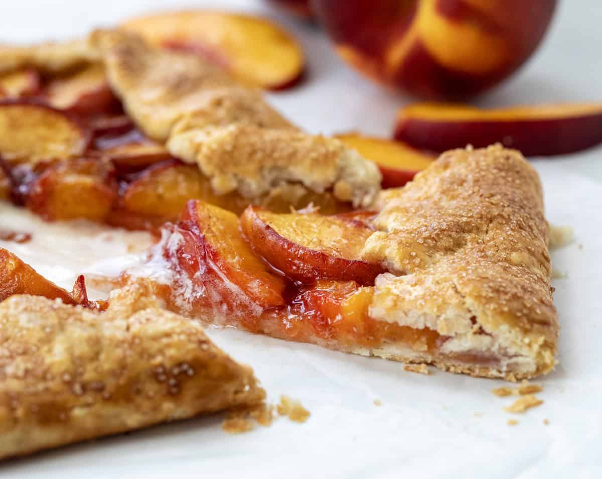 This easy Peach Galette is the perfect summer dessert, made with juicy, fresh peaches wrapped in a buttery, flaky pie crust and baked until golden and bubbling. It is simpler than pie, made with ingredients you probably already have on hand, but just as delicious! Close up of a slice of Peach Galette on a white table.