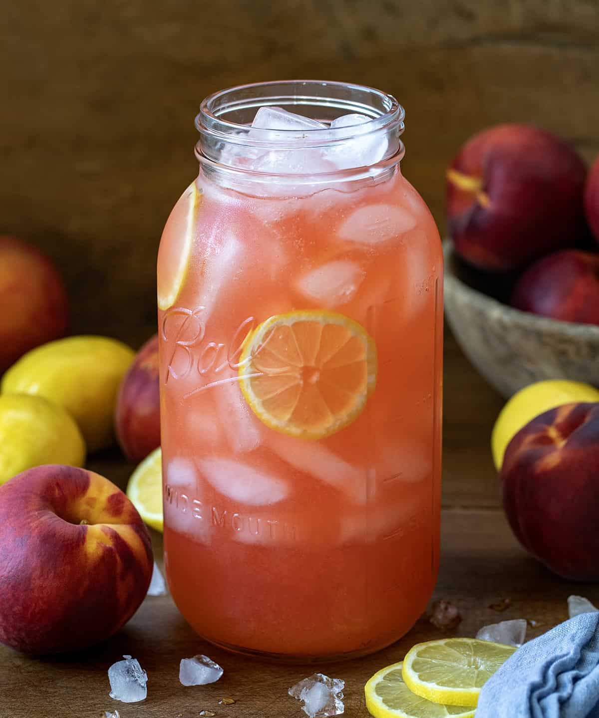 Giant glass jar of Peach Lemonade on a wooden table with fresh peaches and lemons.