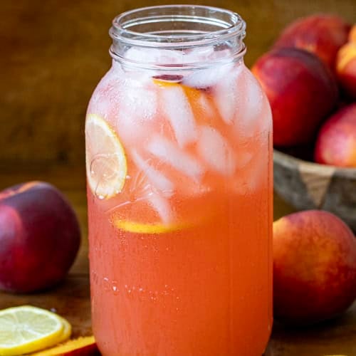Giant glass jar of Peach Lemonade on a wooden table with fresh peaches and lemons.