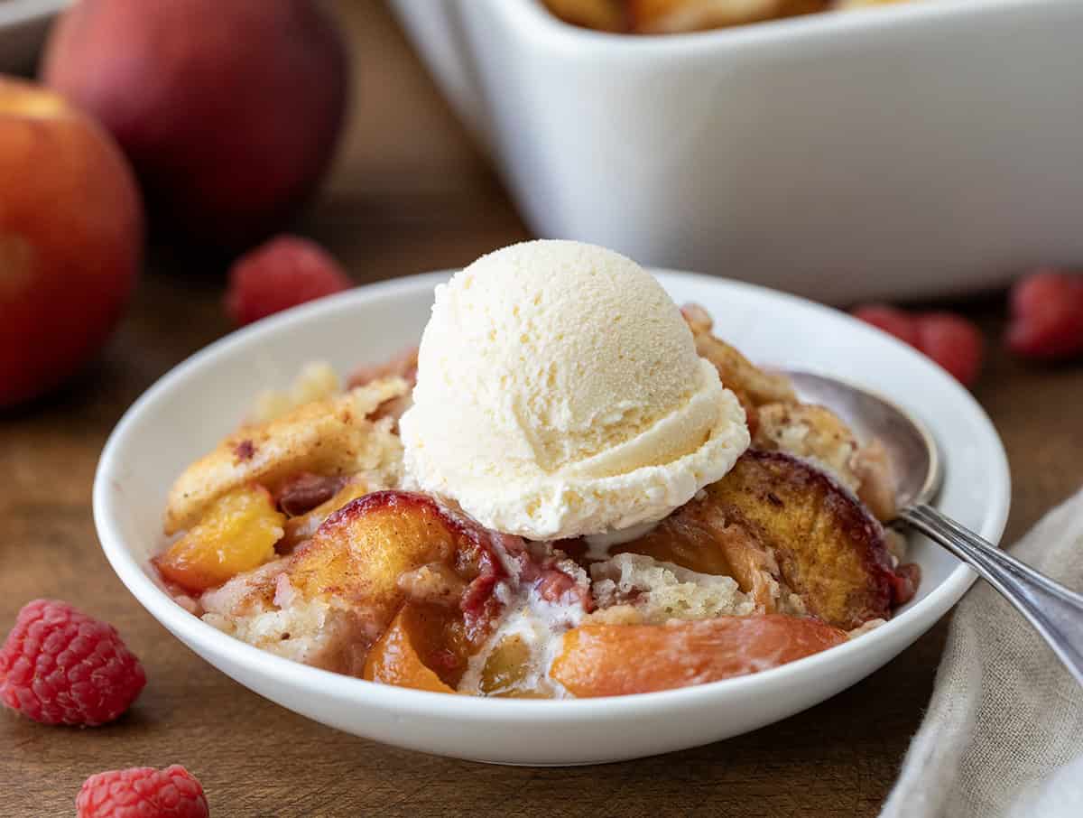 This Raspberry Peach Cobbler is one of my favorite ways to use fresh summer fruit! Juicy peaches and sweet-tart raspberries get tossed with a little cinnamon and nutmeg, then baked under a soft, buttery batter that puffs up and turns golden as it bakes. The fruit gets all bubbly and jammy, and the edges of the cobbler develop that irresistible crisp-chewy texture I love! White plate with Raspberry Peach Cobbler and a melting scoop of vanilla ice cream close up.