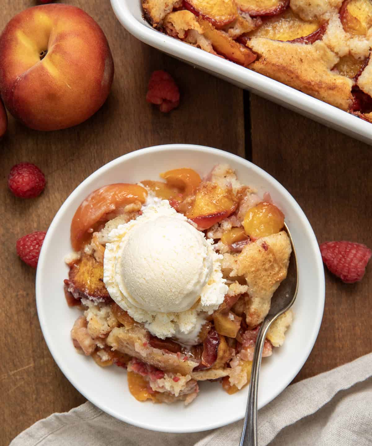 This Raspberry Peach Cobbler is one of my favorite ways to use fresh summer fruit! Juicy peaches and sweet-tart raspberries get tossed with a little cinnamon and nutmeg, then baked under a soft, buttery batter that puffs up and turns golden as it bakes. The fruit gets all bubbly and jammy, and the edges of the cobbler develop that irresistible crisp-chewy texture I love! White plate with Raspberry Peach Cobbler on it and a scoop of ice cream and fresh peaches from overhead.