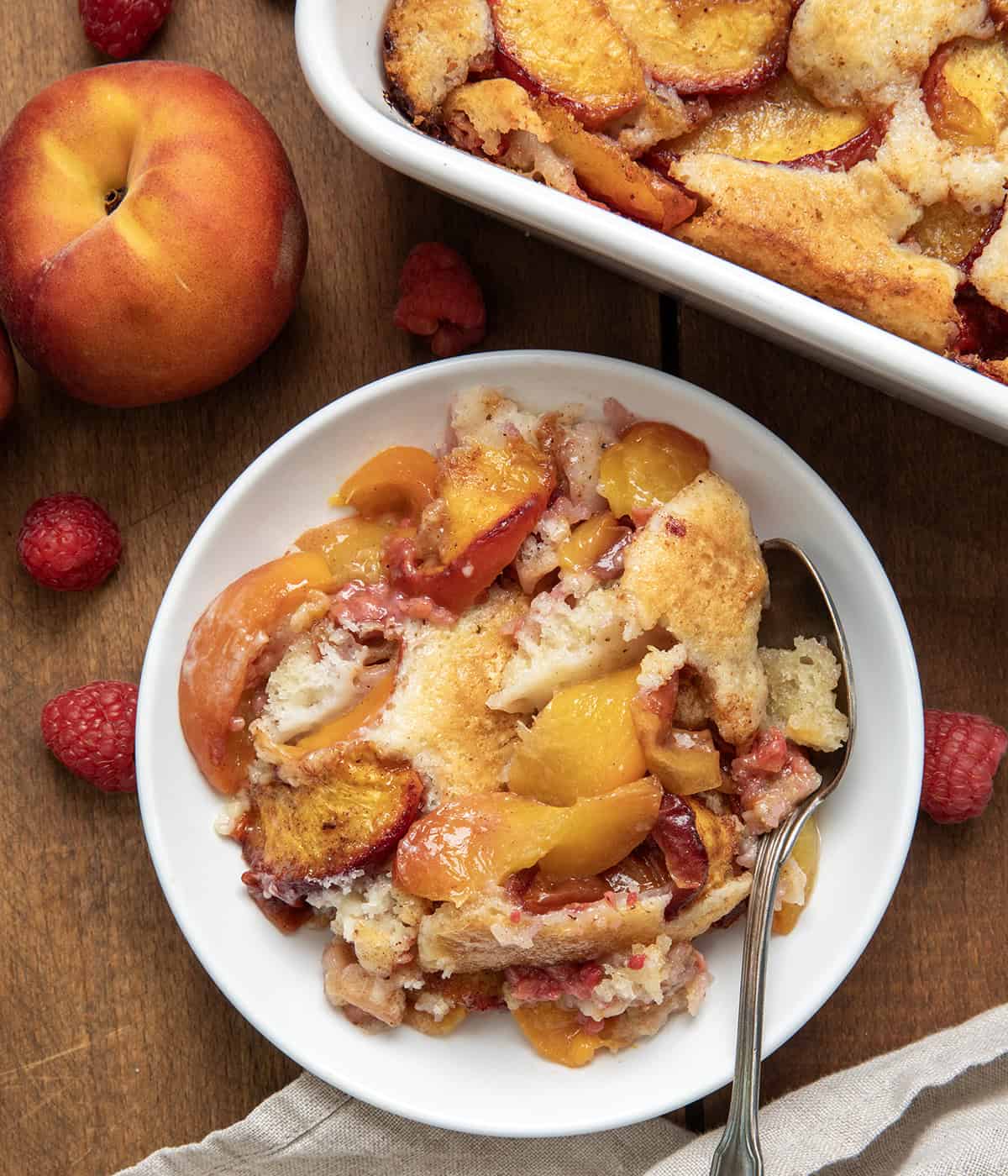 This Raspberry Peach Cobbler is one of my favorite ways to use fresh summer fruit! Juicy peaches and sweet-tart raspberries get tossed with a little cinnamon and nutmeg, then baked under a soft, buttery batter that puffs up and turns golden as it bakes. The fruit gets all bubbly and jammy, and the edges of the cobbler develop that irresistible crisp-chewy texture I love! White plate with Raspberry Peach Cobbler and a spoon on a wooden table from overhead.