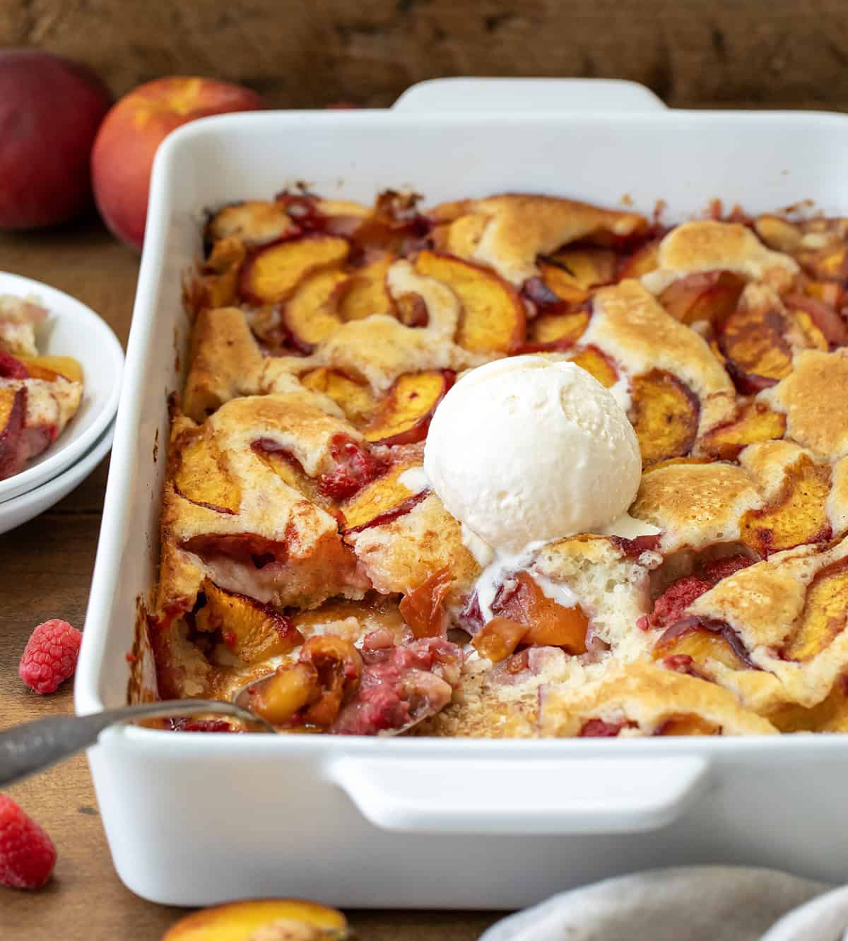 This Raspberry Peach Cobbler is one of my favorite ways to use fresh summer fruit! Juicy peaches and sweet-tart raspberries get tossed with a little cinnamon and nutmeg, then baked under a soft, buttery batter that puffs up and turns golden as it bakes. The fruit gets all bubbly and jammy, and the edges of the cobbler develop that irresistible crisp-chewy texture I love! Pan of Raspberry Peach Cobbler with ice cream on top, a portion removed, and a spoon resting on the pan.