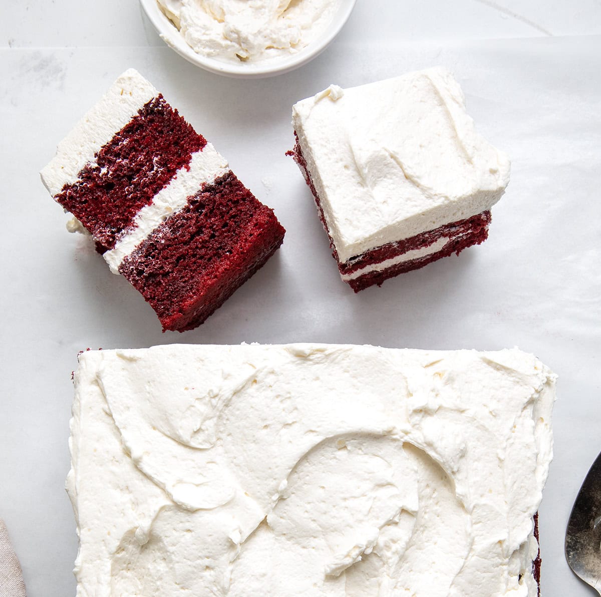 If you love red velvet, this Sheet Pan Red Velvet Cake is the easy, crowd-friendly dessert you’ve been looking for. It’s soft, moist, and full of that classic cocoa-vanilla flavor. The cake is layered with silky ermine frosting for a beautiful stacked presentation that feels extra special, without the stress of baking two separate layers! Sheet Pan Red Velvet Cake on a white table with two slices in front, one upright and one on its side.