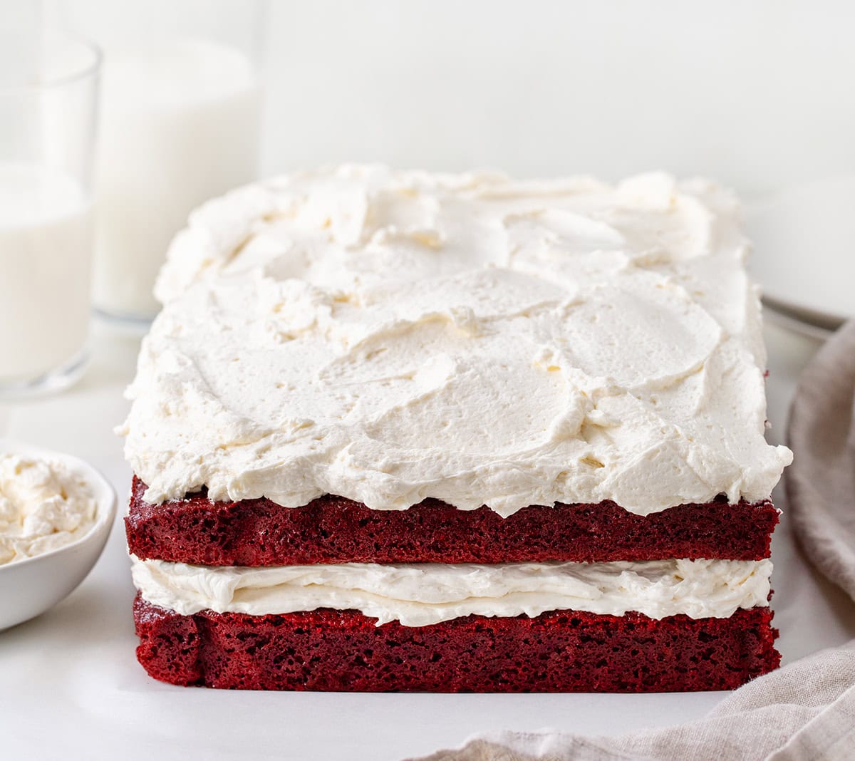 If you love red velvet, this Sheet Pan Red Velvet Cake is the easy, crowd-friendly dessert you’ve been looking for. It’s soft, moist, and full of that classic cocoa-vanilla flavor. The cake is layered with silky ermine frosting for a beautiful stacked presentation that feels extra special, without the stress of baking two separate layers! Sheet Pan Red Velvet Cake on a white table with glasses of milk.