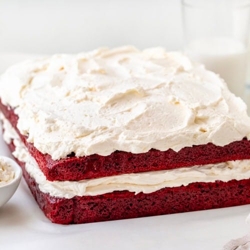 Sheet Pan Red Velvet Cake on a white table with ermine frosting and glasses of milk.
