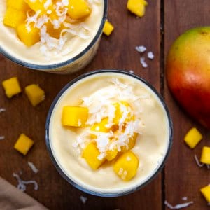 Two Tropical Mango Milkshakes on a wooden table with fresh mango and coconut.