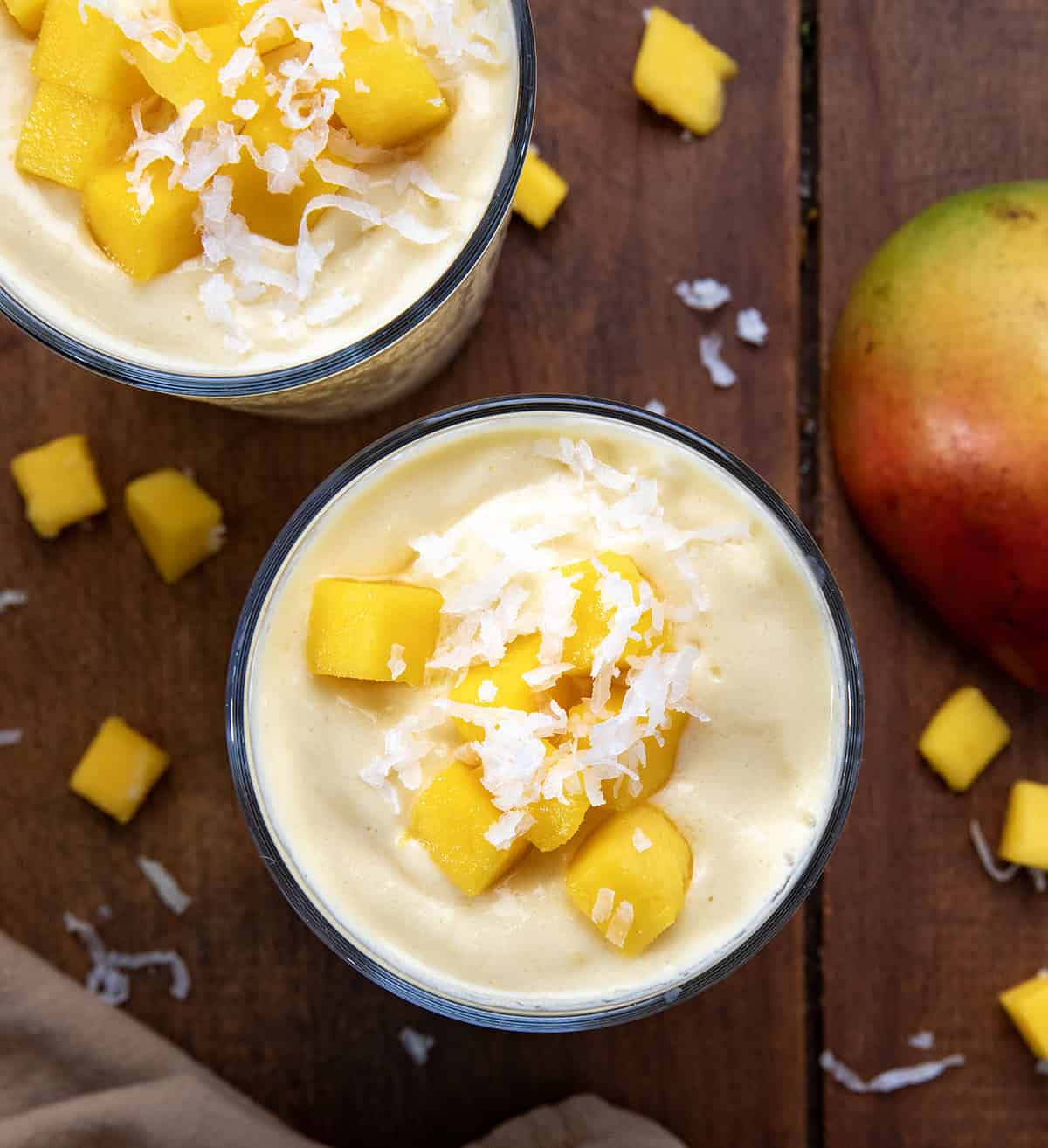Two Tropical Mango Milkshakes on a wooden table with fresh mango and coconut.