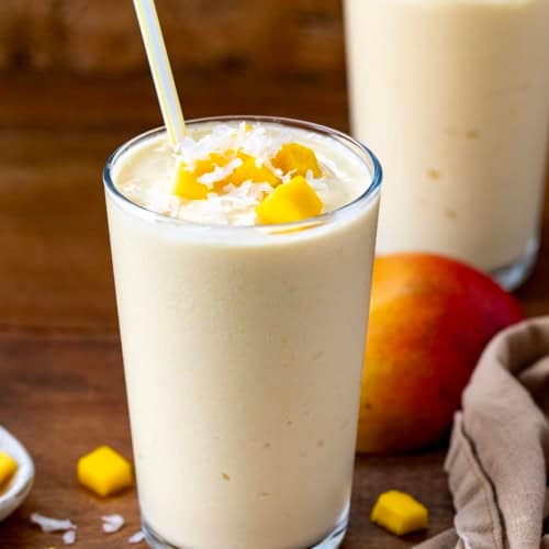 Two Tropical Mango Milkshakes on a wooden table with fresh mango and coconut.