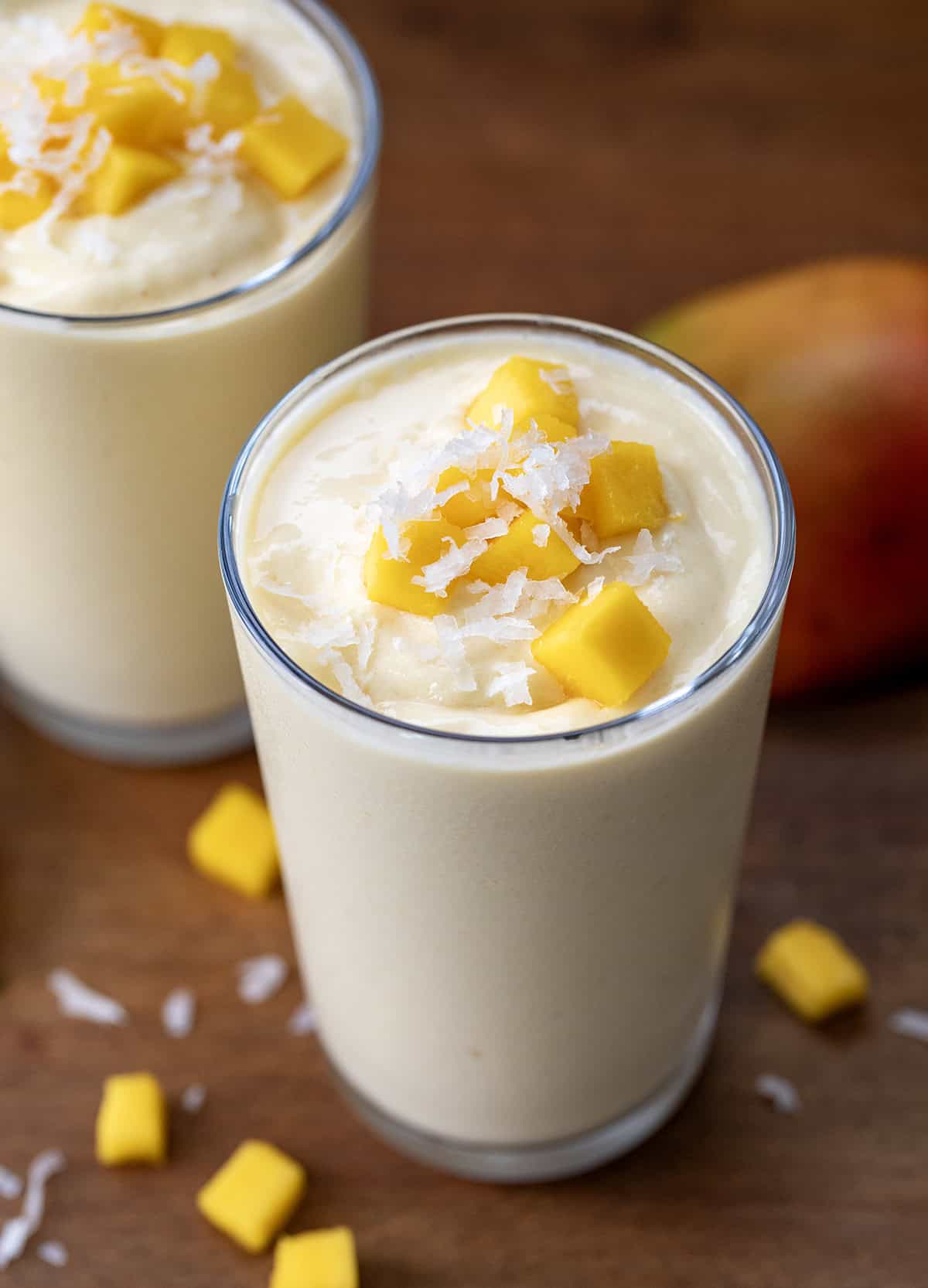 Two Tropical Mango Milkshakes on a wooden table with fresh mango and coconut.