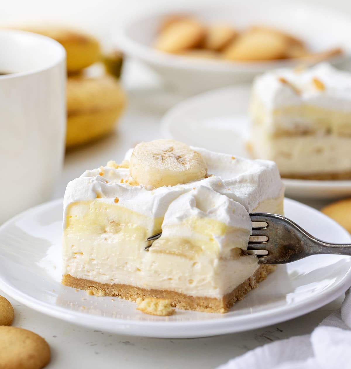 These Banana Cream Bars are an easy summer dessert that everyone will love! The no-bake dessert has a vanilla wafer crust, creamy filling, fresh bananas, and fluffy topping. Itโs cool, creamy, and perfect for hot days when you donโt want to turn on the oven. Fork taking a piece of a Banana Cream Pie Bar on a white plate.