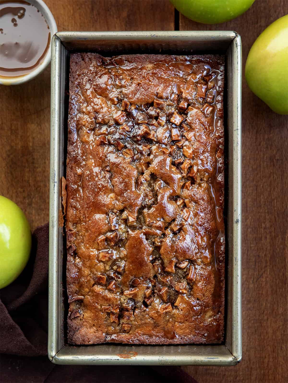 Caramel Apple Fritter Bread takes my fan-favorite Apple Fritter Bread and layers in a sweet caramel drizzle for the ultimate fall baking treat. Soft and tender with cinnamon-sugar apples swirled throughout, this bread is like biting into a caramel apple fritter, with no frying required! Pan of Caramel Apple Fritter Bread on a wooden table with green apples from overhead.