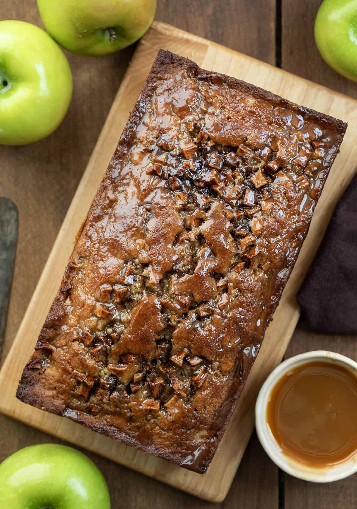 Caramel Apple Fritter Bread takes my fan-favorite Apple Fritter Bread and layers in a sweet caramel drizzle for the ultimate fall baking treat. Soft and tender with cinnamon-sugar apples swirled throughout, this bread is like biting into a caramel apple fritter, with no frying required! Caramel Apple Fritter Bread on a cutting board on a wooden table with caramel and apples from overhead.