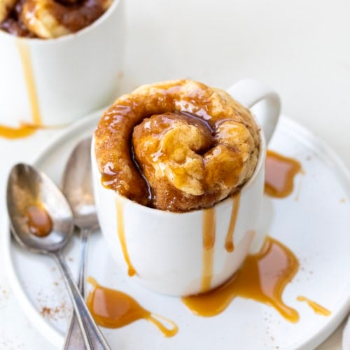 Caramel Roll Mug Cake in a white mug on a white table with spoons.