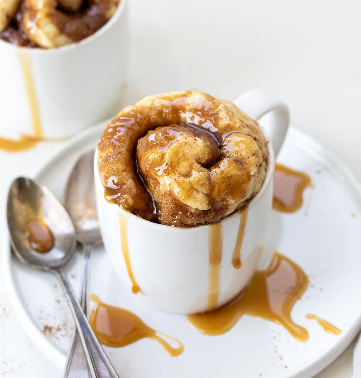 Caramel Roll Mug Cake in a white mug on a white table with spoons.